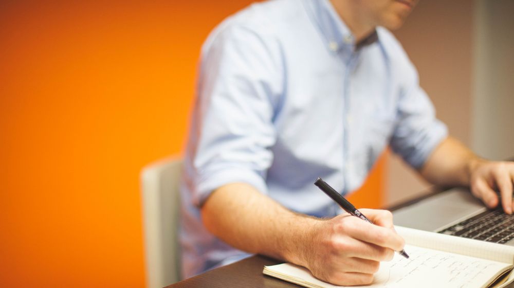 Man writing on notepad while working on laptop, writing questions for an exam.